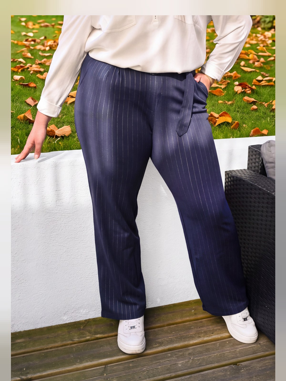Pantalon marine à fines rayures blanches, coupe droite, porté avec des baskets blanches sur une terrasse en bois
