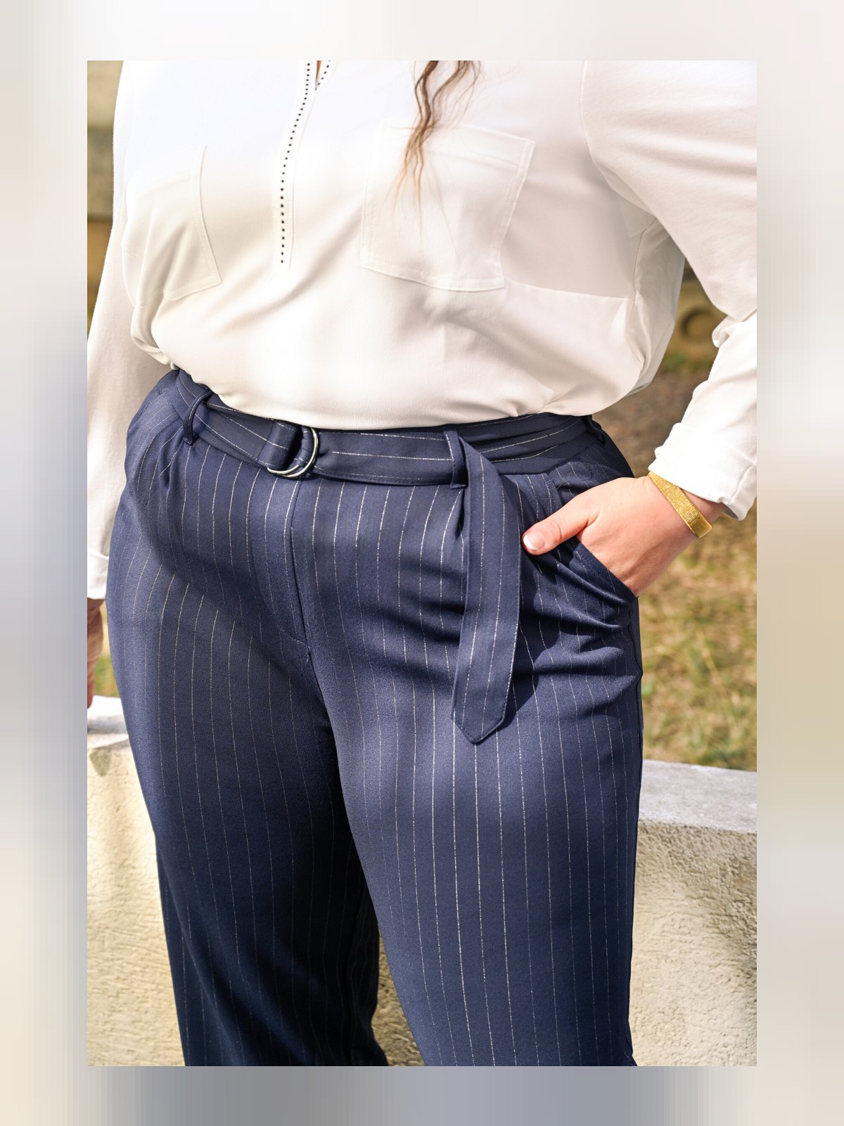 Gros plan sur la ceinture à boucle et les fines rayures blanches du pantalon marine, main dans la poche