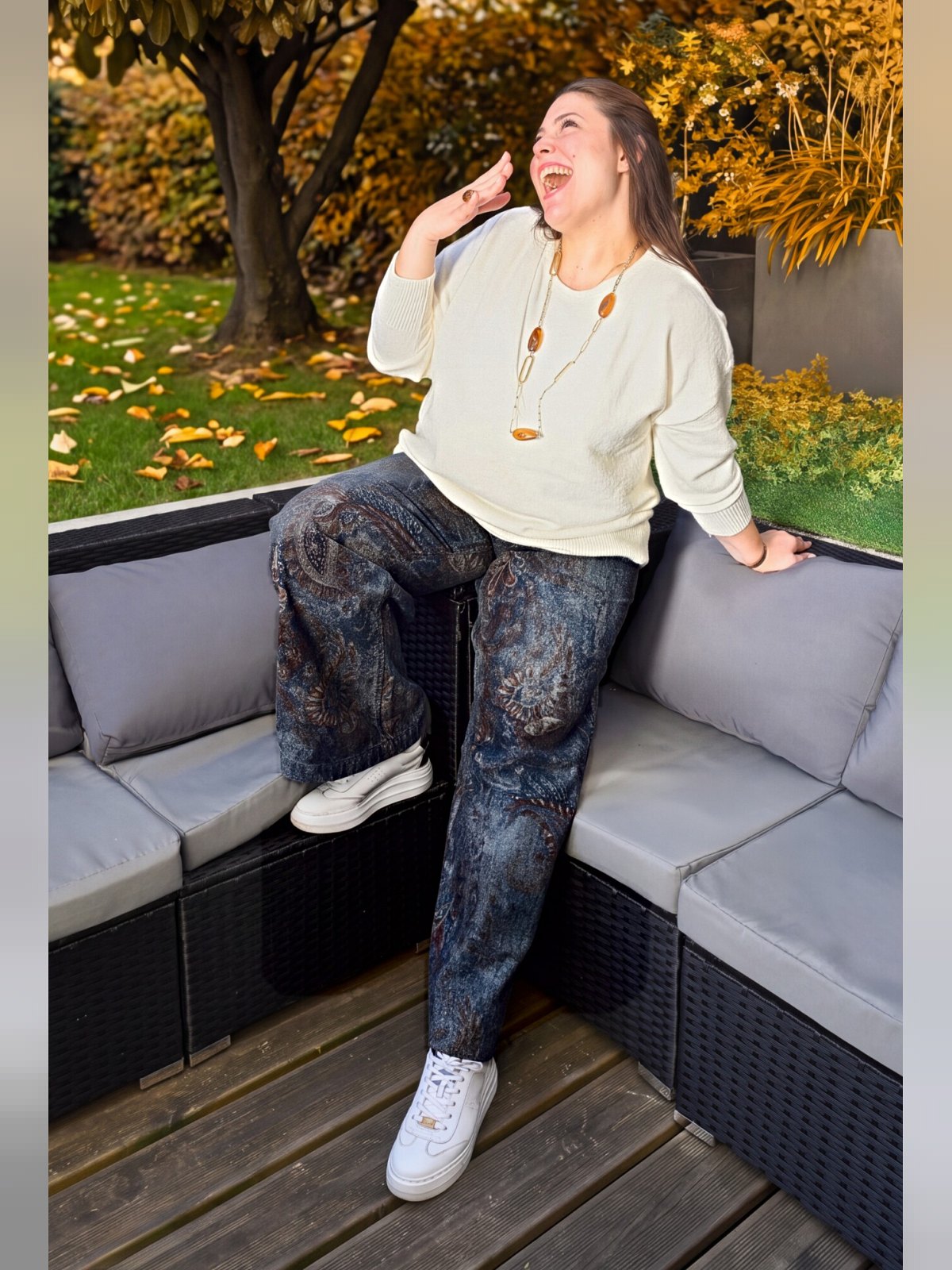 Pull écru porté par une femme assise sur un salon de jardin, avec pantalon imprimé et baskets blanches